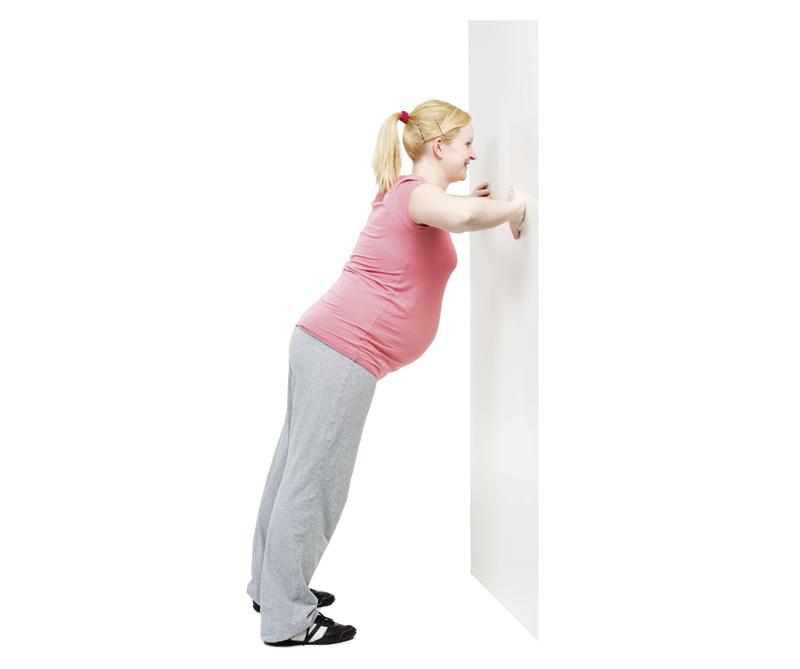 A pregnant woman is leaning towards a wall to make a chest muscle exercise.