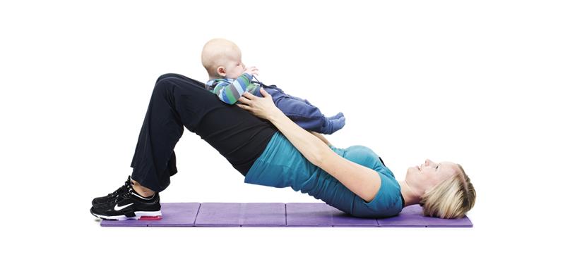 A woman is lifting her pelvis into a bridge position while holding her baby on thighs.