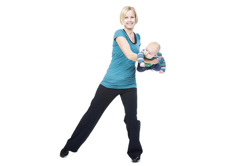A woman is holding her baby while doing a side tap exercise.