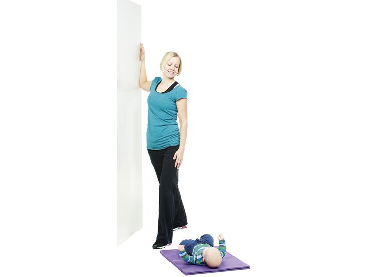 A woman is stretching the chest muscles against a wall while her baby is lying on a mat.